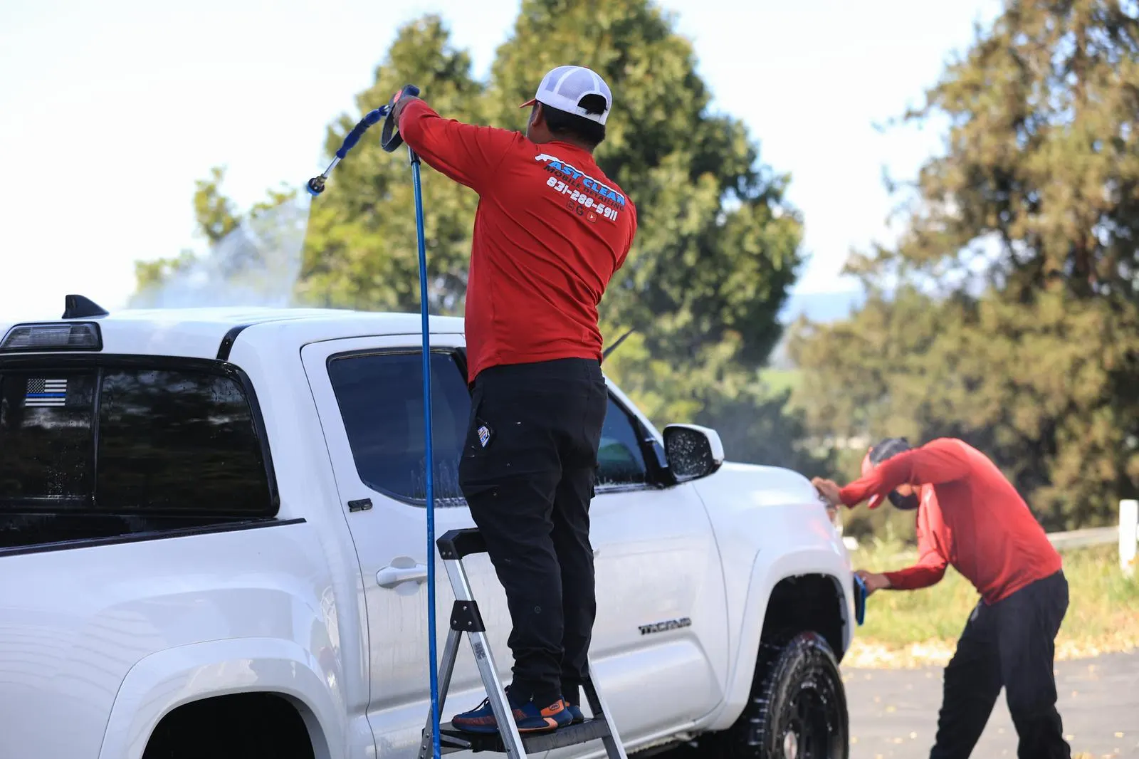 Two Fast Clean Mobile Detailing technicians washing a white Toyota Tacoma truck in a driveway, one on a stepladder rinsing the roof.