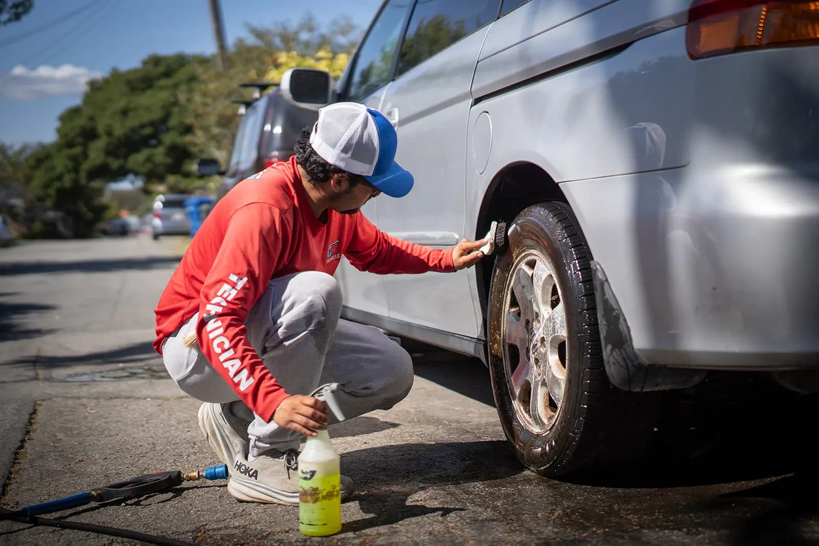 Fast Clean technician kneeling to scrub a wheel and fender on a silver minivan parked on a residential street.