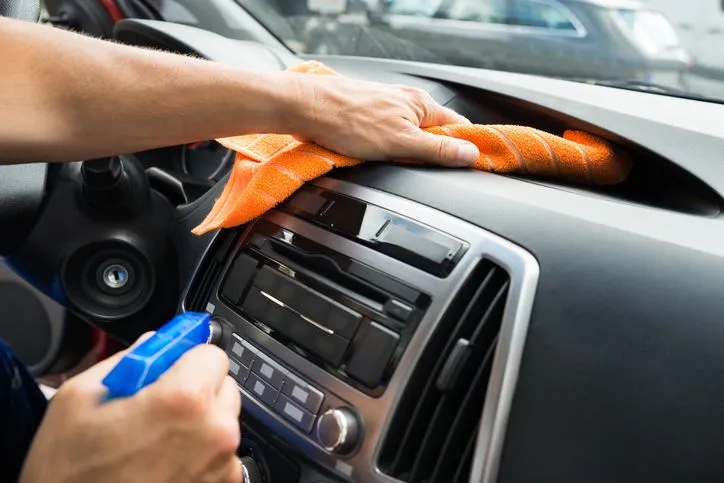 Detailer wiping down a car dashboard with an orange microfiber cloth and cleaning spray.